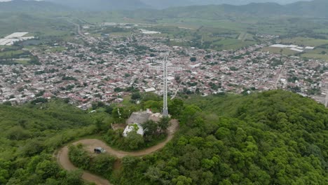 Aerial-orbiting-shot-of-cross-on-green-hilltop-with-city-of-Tecalitlan-in-Valley-and-green-landscape-during-foggy-day