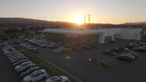 Beautiful-sun-setting-behind-a-mountain,-over-a-BMW-dealership-lot-full-of-cars,-aerial-rising