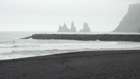 Islands-Schwarzer-Sandstrand-Mit-Rauen-Wellen-Und-Fernen-Felsformationen-Unter-Einem-Nebligen,-Bewölkten-Himmel
