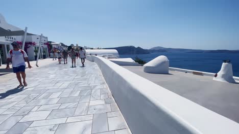 People-On-White-Sidewalk-Near-Cafe-|-Oia-Santorini-Greece-Greek-Island-in-Aegean-Sea,-Travel-Tourist-Vacation-Immersive-Moving-Walk-Along-Crowds-Shopping-in-White-Marble-Cliffside-and-City,-Europe,-4K