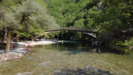 Tourists-marvel-at-the-breathtaking-scenery-on-the-riverbank-of-the-Voidomatis-River,-with-the-iconic-Aristi-Bridge-adding-to-the-beauty-of-the-landscape
