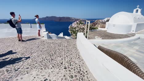 Tourists-Watching-Peoples-Posing-|-Oia-Santorini-Greece-Greek-Island-in-Aegean-Sea,-Travel-Tourist-Vacation-Immersive-Moving-Walk-Along-Crowds-Shopping-in-White-Marble-Cliffside-and-City,-Europe,-4K