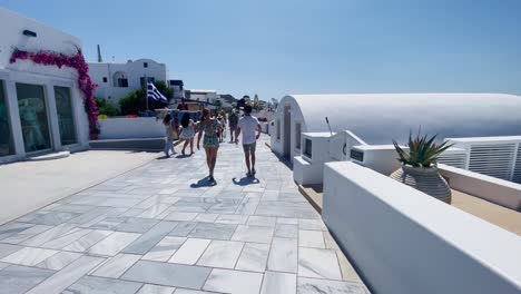 Gente-Caminando-En-La-Acera-Blanca-|-Oia-Santorini-Grecia-Isla-Griega-En-El-Mar-Egeo,-Viajes-Turísticos-Vacaciones-Inmersivas-Caminatas-En-Movimiento-Junto-A-Multitudes-De-Compras-En-Acantilados-De-Mármol-Blanco-Y-Ciudad,-Europa,-4k