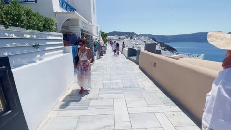 Couple-Walking-Near-Wide-View-of-Sea-|-Oia-Santorini-Greece-Greek-Island-in-Aegean-Sea,-Travel-Tourist-Vacation-Immersive-Moving-Walk-Along-Crowds-Shopping-in-White-Marble-Cliffside-and-City,-Europe4K