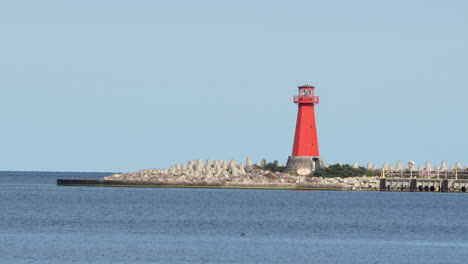 :-A-vibrant-red-lighthouse-stands-on-a-rocky-outcrop-at-the-edge-of-the-sea-under-a-clear-sky