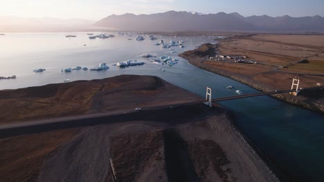 Serena-Laguna-Glacial-Con-Icebergs,-Un-Puente-Que-Cruza-Y-Un-Telón-De-Fondo-De-Montañas-Al-Atardecer-Desde-Una-Perspectiva-Aérea-En-Islandia