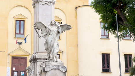 A-marble-angel-statue-with-spread-wings-stands-in-front-of-a-historic-building,-symbolizing-peace-or-remembrance,-with-Italian-flags