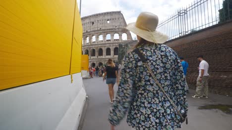 Rome-Immersive-POV:-Moving-In-Busy-Streets-to-Chiesa-Santi-Luca-e-Martina,-Italy,-Europe,-Walking,-Shaky,-4K-|-Woman-In-Hat-Walking-Near-Long-Line-Of-Tourists-By-Sign-In-Front-Of-Colosseum