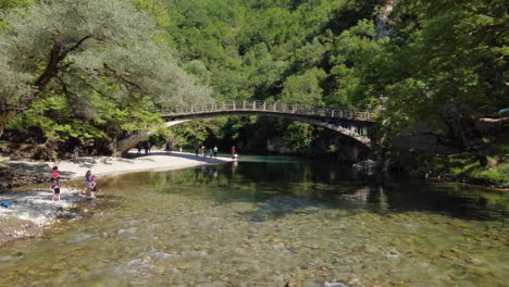 Tourists-enjoy-the-scenery-on-the-banks-of-the-Voidomatis-River,-where-the-famous-Aristi-Bridge-adds-to-the-beauty-of-the-landscape