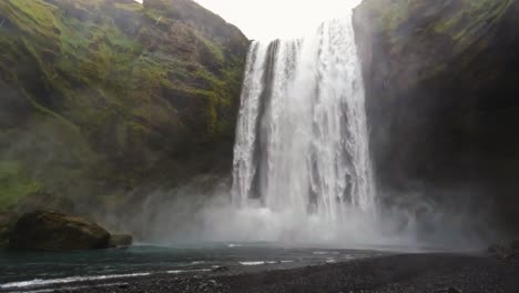 Majestätischer-Wasserfall,-Der-Moosige-Klippen-Hinabstürzt-Und-In-Ein-Ruhiges-Becken-Stürzt,-In-Dem-Nebel-über-Dem-Schwarzen-Kiesstrand-In-Island-Aufsteigt
