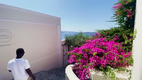 Employee-Near-Pink-and-White-Flower-|-Oia-Santorini-Greece-Greek-Island-in-Aegean-Sea,-Travel-Tourist-Vacation-Immersive-Moving-Walk-Along-Crowds-Shopping-in-White-Marble-Cliffside-and-City,-Europe,4K