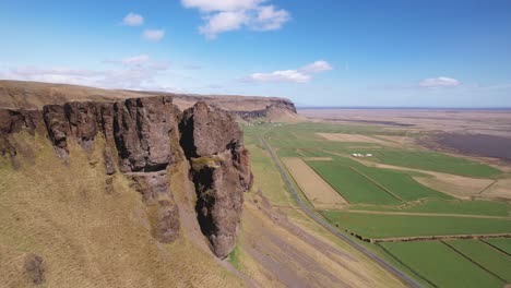 Rugged-cliff-edge-overlooking-vast-green-agricultural-fields-under-a-clear-blue-sky-in-Iceland,-aerial-view