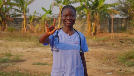 Doctora-Negra-Africana-Te-Amo-Con-Gesto-De-Manos-Frente-A-La-Cámara-Sonriendo-En-Una-Remota-Clínica-Hospitalaria