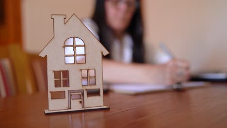 Professional-Woman-Finishing-Paperwork-in-Blurry-Background-With-Focus-On-Miniature-House-Icon-On-Office-Table