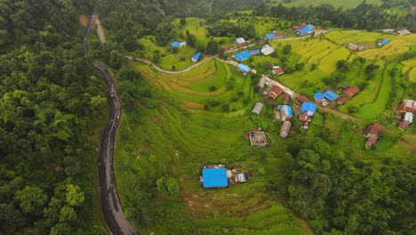 Aerial-top-down-shot-of-car-on-road-in-jungle-of-Nepal,-showing-colorful-houses-between-plantaiton-fields-at-hillside---Tilt-up