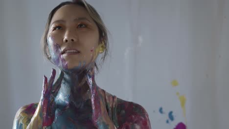 Asian-girl-covered-in-colorful-paint-poses-in-studio,-handheld-closeup