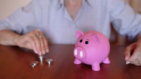 Woman-Putting-Coins-Into-Pink-Piggy-Bank-On-Table