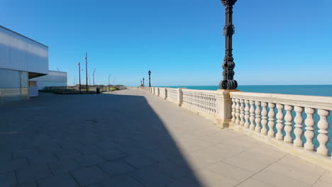 Eine-Breite-Strandpromenade-In-Cádiz-Mit-Einer-Kunstvollen-Straßenlaterne-Und-Einer-Klassischen-Balustrade,-Die-Einen-Weiten-Blick-Auf-Das-Meer-Unter-Einem-Klaren-Blauen-Himmel-Bietet