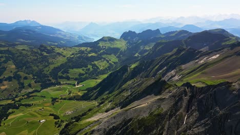 View-from-a-high-peak,-overlooking-a-village-with-large-mountain-range-in-the-background,-surrounded-in-green-landscape
