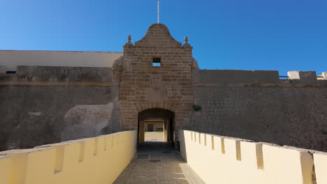 A-stone-gateway-at-the-entrance-of-an-ancient-fortress-in-Cádiz,-with-a-path-leading-through-it,-flanked-by-high-walls-under-a-clear-blue-sky