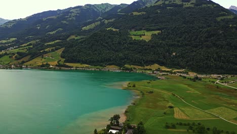 Panoramic-view-of-a-lakefront-village,-lush-green-field,-pristine-lake,-surrounded-by-large-tree-covered-mountains