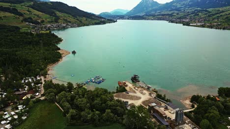 Aerial-view-of-a-pristine-lake,-with-a-dock-and-boats,-on-the-edge-of-a-village,-surrounded-by-mountains