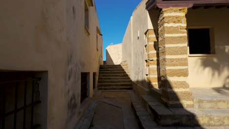 A-narrow-alleyway-in-Cádiz-with-traditional-stone-stairs-leading-up-between-old-buildings,-cast-in-the-golden-light-of-the-sun