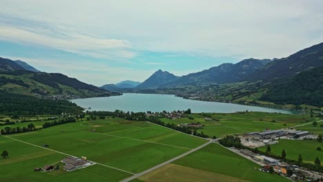 Aerial-view-of-a-remote-lakefront-village,-surrounded-by-lush-green-fields-and-mountains,-with-a-high-peak-mountaintop-in-the-distance