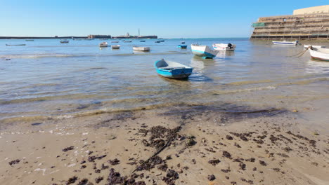 Shallow-waters-edge-on-a-sandy-beach-in-Cádiz-with-several-small-boats-floating-near-the-shore,-clear-blue-sky-above,-and-a-distant-view-of-the-city's-maritime-structures