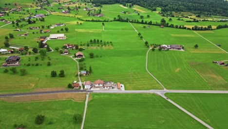Luftaufnahme-Eines-Kleinen-Abgelegenen-Dorfes,-In-Einem-Großen-Grünen-Feld,-An-Einem-Berghang-In-Den-Schweizer-Alpen