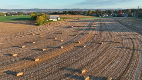 Aerial-view-of-a-farmland-with-neat-furrows,-scattered-hay-bales,-and-a-tractor-at-sunset