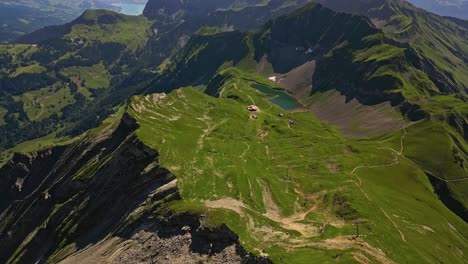 Aerial-view-of-a-mountain-range-in-summer-with-green-grass,-peaks-and-slopes-and-a-little-pond,-with-a-lake-and-mountains-in-the-background