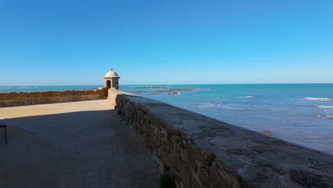 Un-Paseo-Costero-En-Cádiz-Con-Una-Pequeña-Garita-Al-Final-De-Un-Muro-De-Piedra,-Con-Vistas-Al-Océano-Atlántico-Bajo-Un-Vasto-Cielo-Azul.