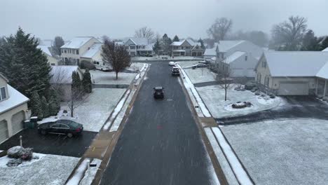 Aerial-tracking-shot-of-car-driving-through-snow-covered-neighborhood-during-snow-flurries-in-winter