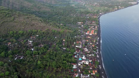 Drone-top-down-view-of-a-small-village-stretching-out-along-the-coast-with-black-sand-beaches-and-volcano-in-the-background