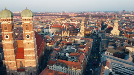 Aerial-passing-near-the-towers-of-the-architectural-marvel,-Munich-Frauenkirche,-Rathaus-the-aerial-perspective-unveils-the-cityscape-of-Munich-on-the-horizon-during-low-angle-sunshine
