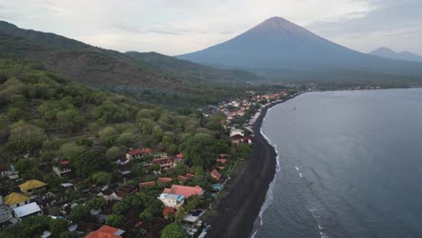 Drone-flying-over-a-small-coastal-village-with-black-sand-beaches-and-volcano-in-the-background