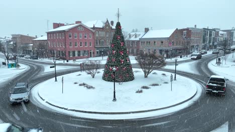 Snowy-town-square-with-decorated-Christmas-tree-in-traffic-roundabout,-surrounded-by-historic-buildings
