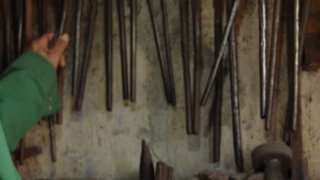 A-blacksmith-selects-a-pair-of-tongs-from-his-workshop's-wall,-test-them-on-a-piece-of-metal