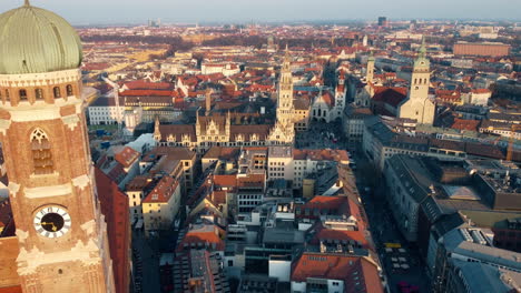 Aerial-passing-near-towers-of-architectural-marvel-of-Munich-Frauenkirche,-revealing-Munich-city-landscape-on-horizon