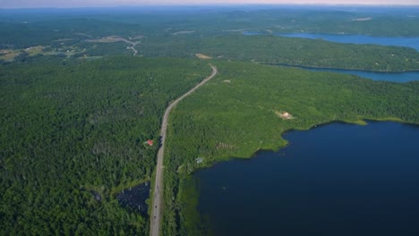 Aerial-wide-shot-of-cars-passing-on-Canada's-highway-next-to-lush-trees-and-clear-blue-lakes