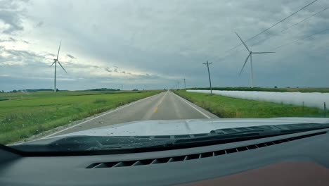 POV---driving-on-country-road-past-hay-fields-with-wrapped-bales,-corn-fields-and-wind-turbines