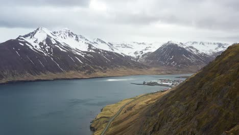 Vista-Aérea-De-Un-Sereno-Fiordo-Islandés-Con-Montañas-Nevadas-Y-Un-Pueblo-Costero