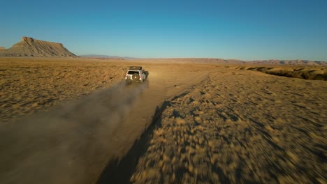 Exhilarating-FPV-drone-flight-tracks-white-SUV-driving-on-dusty-desert-road-toward-Factory-Butte,-Utah