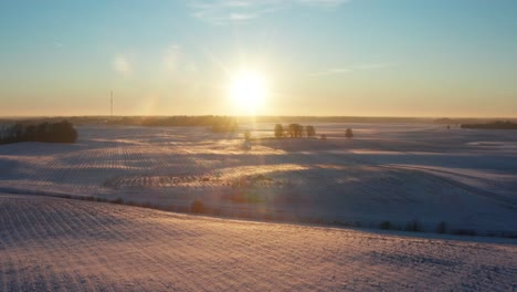 Filmische-Luftaufnahme-Eines-Hellen-Wintersonnenuntergangs-über-Einem-Verschneiten-Landwirtschaftlichen-Feld