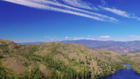 Drone-shot-panning-right-then-pulling-away-revealing-a-stunning-landscape-of-trees,-mountains-and-a-lake-on-a-bright-sunny-day