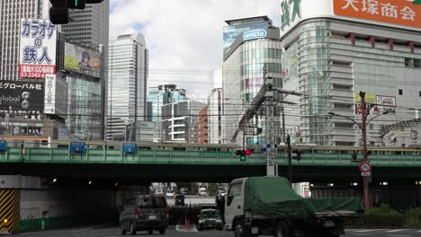 Bezirk-Shinjuku,-Autos-Fahren-Unter-Der-Eisenbahnbrücke-Hindurch,-Vorbeifahrende-Züge,-Geschäftsgebäude-Im-Hintergrund
