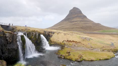 Majestuosa-Cascada-De-Kirkjufellsfoss-Con-La-Montaña-De-Kirkjufell-Al-Fondo,-Los-Visitantes-Exploran-El-Pintoresco-Paisaje-Islandés