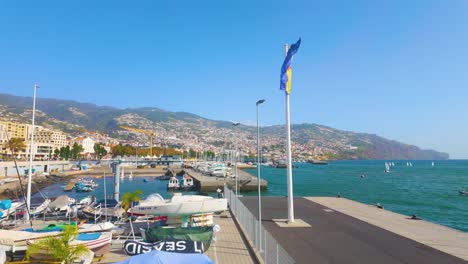Waving-Flag-of-Funchal-in-Portugal-during-sunny-day-at-harbor-with-beach-and-town-on-mountain-slope---wide-shot