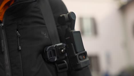 Close-up-of-Osmo-Pocket-3-camera-attached-to-backpack-on-man's-chest,-turns-on-with-press-of-button